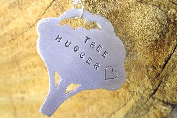 Pendant shaped like a tree on wood background, text reads "TREE HUGGER" with a heart.